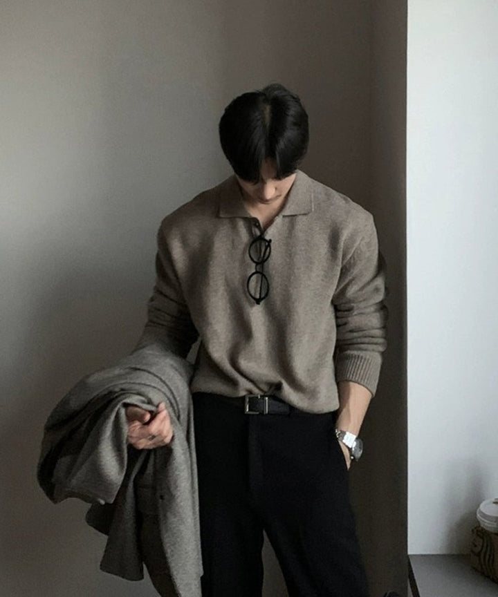 壁際にスッと立ち、アウターを片手に持って少し俯く男性のフロントカット。ドロップショルダーの滑らかなラインと、タックインした際の美しいブラウジングがよくわかる画像。