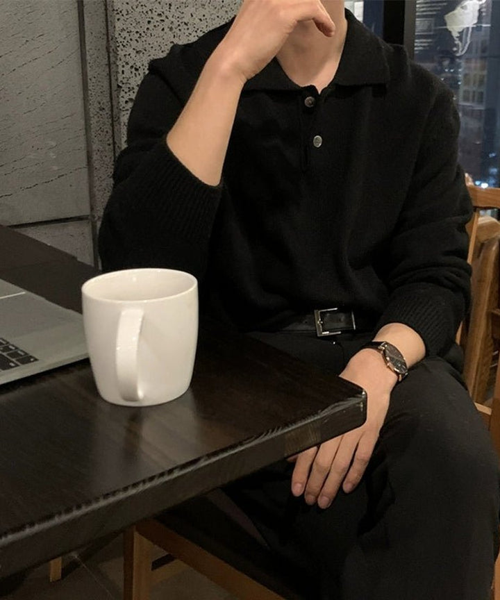 カフェやオフィスのテーブル席に座り、手元に白いマグカップを置いた男性のシチュエーションカット。シックな空間に溶け込む黒ニットの落ち着いた表情を演出した画像。