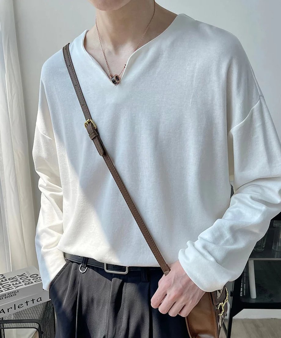 首元に浅いV字のカッティングが入ったホワイトのVネック長袖カットソーを着用し、茶色のレザーショルダーストラップを斜め掛けして手で持つ男性の斜め前からの写真。