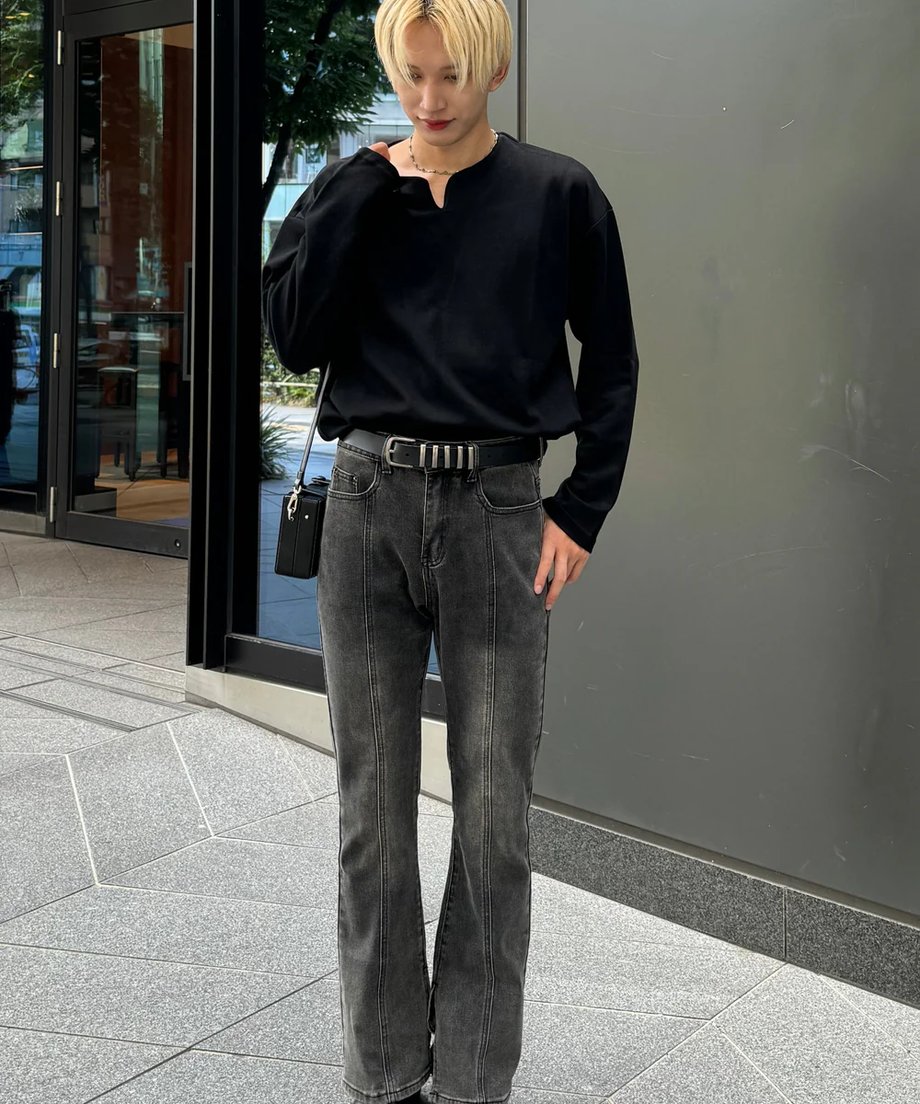 屋外で、黒のVネック長袖カットソーにセンタープレスの入ったグレーのウォッシュドフレアデニム、黒のベルトとショルダーバッグを合わせた金髪の男性の全身コーディネート写真。