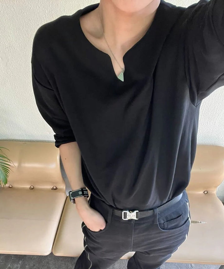 ベージュのソファの前で、黒のVネック長袖Tシャツに黒のパンツとデザインベルトを合わせ、左手をポケットに入れた男性の俯瞰写真。