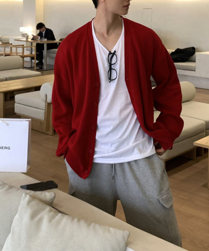 レッドのオーバーサイズカーディガンを羽織り、両手をポケットに入れてリラックスして佇む男性のスタイリングカット。インナーの白Tシャツに黒縁メガネが掛けられている。