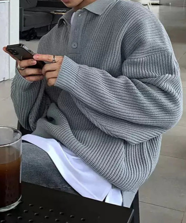 グレーのポロカラーリブニットを着用し、カフェのテラス席でスマートフォンを操作している男性のサイドカット。ゆったりとしたボリュームスリーブのシルエットが際立つ一枚。