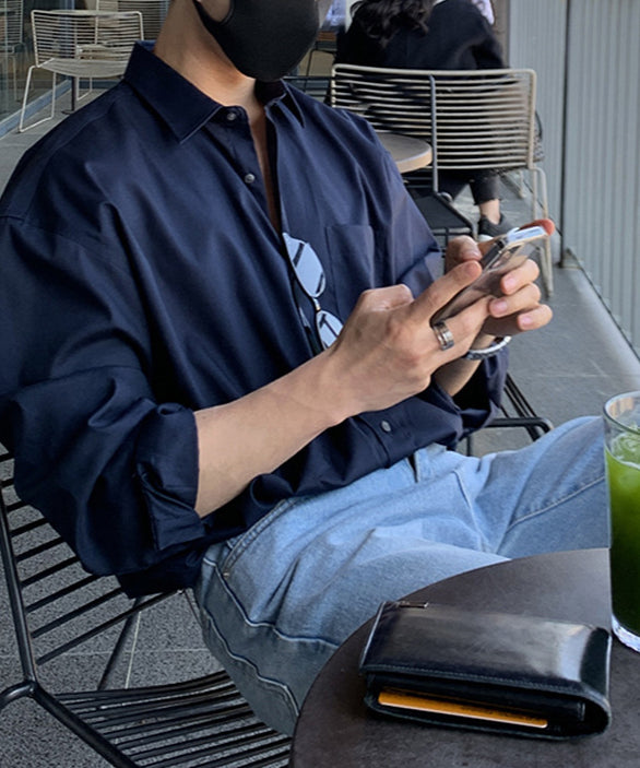 カフェの屋外テラス席で、ネイビーのルーズシャツを着用し、両手でスマートフォンを操作している人物のアップ。シャツのボタンを少し開けて抜け感を出し、ウォッシュデニムを合わせた都会的なダークトーンスタイル。手前のテーブルには黒いレザーの二つ折り財布が置かれている。