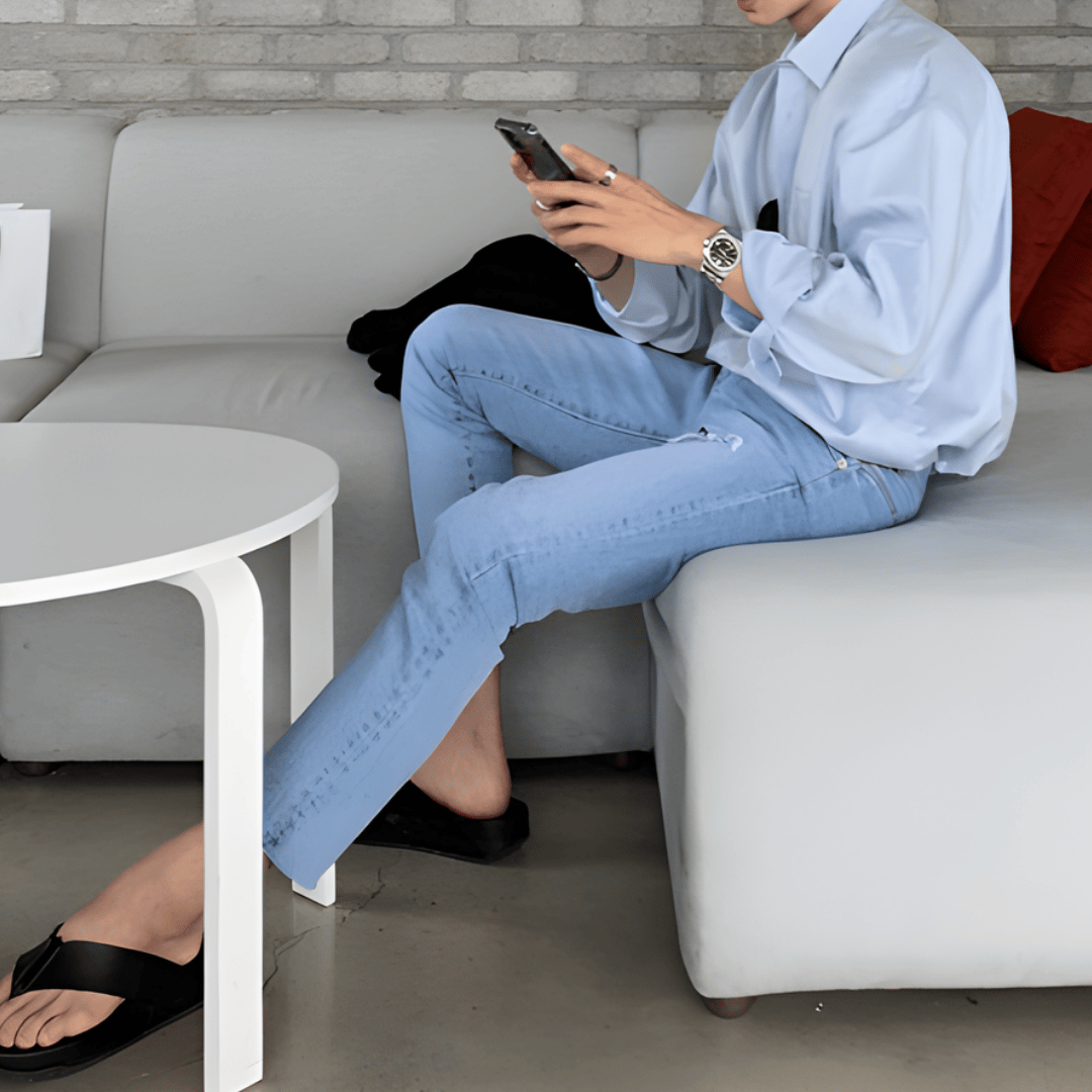 白を基調としたスタイリッシュなカフェのソファ席に座り、両手でスマートフォンを操作している人物のサイドルック。爽やかなライトブルーのシャツに、ダメージ加工が施されたスリムなライトブルーデニム、足元には黒のレザー仕様トングサンダルを合わせた清涼感のあるコーディネート。