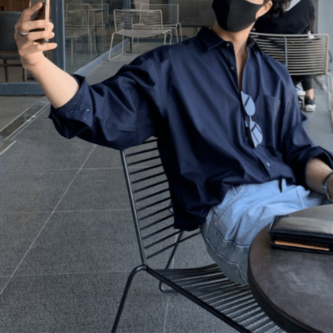 テラス席のワイヤーチェアに少し深く腰掛け、左手でスマートフォンを高く掲げて自撮りや撮影をしているようなポーズの人物。ネイビーのオーバーサイズシャツをフロントインし、ライトブルーのウォッシュデニムを合わせた、こなれ感のあるシティカジュアルスタイルです。