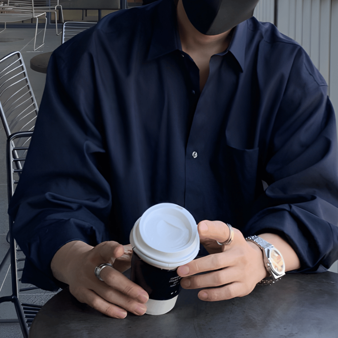 カフェのモダンなテーブル席で、ネイビーのルーズシャツを着用した人物が、両手でテイクアウトカップを包むように持っている手元のアップ。両手の指にはシンプルなシルバーリング、左腕にはメタルバンドの腕時計が光り、ダークトーンのシャツに上品なアクセントを添えています。