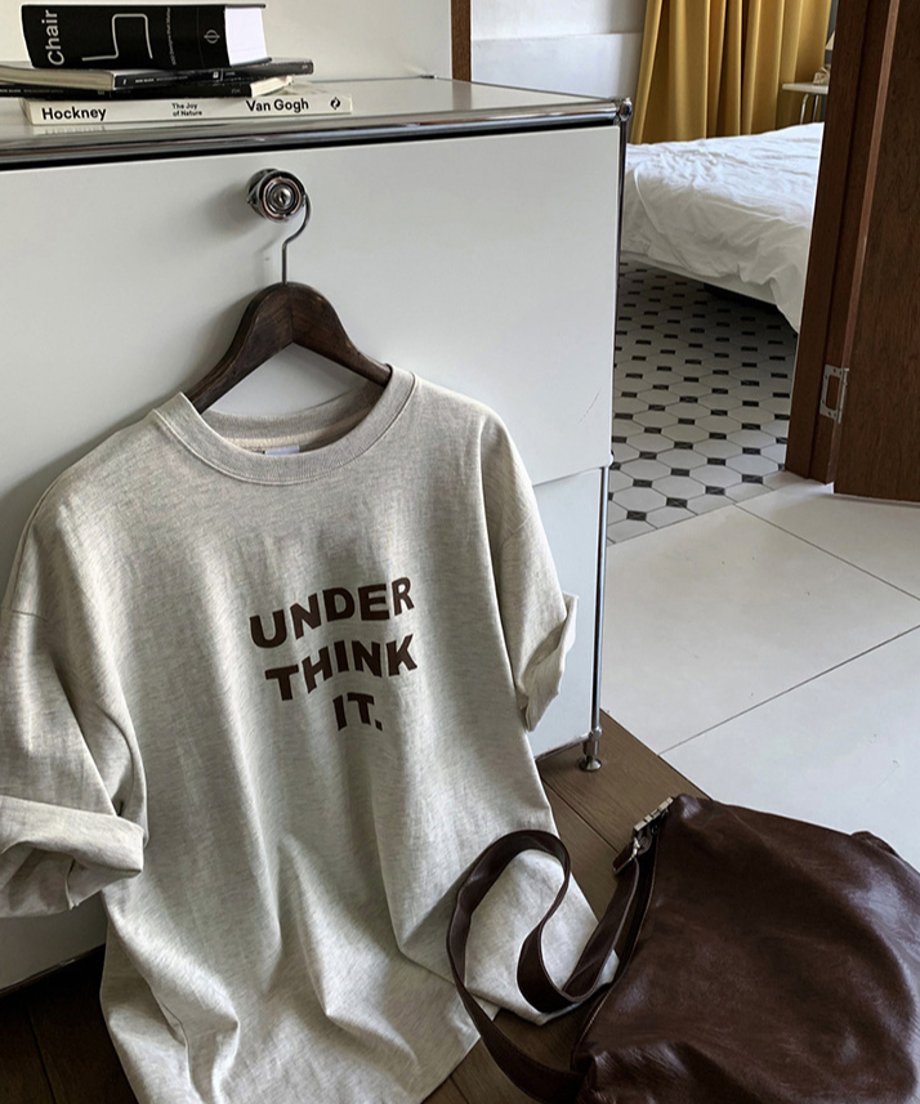 ヴィンテージ調のインテリアの中に、木製ハンガーに掛けられたオートミールのグラフィックTシャツがチェストに立て掛けられているイメージカット。手前にはブラウンのレザーショルダーバッグが置かれている。