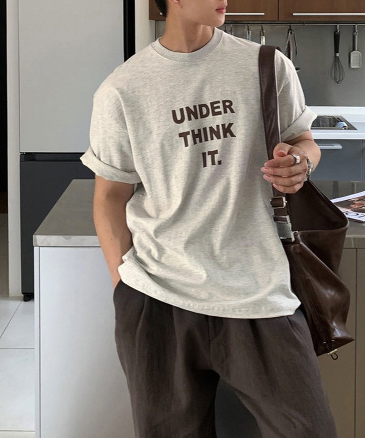 袖口をロールアップしたオートミールのグラフィックTシャツに、ブラウンのパンツとレザーバッグを斜め掛けした男性が、モダンなキッチンカウンターの横に佇むクールなスタイリングカット。