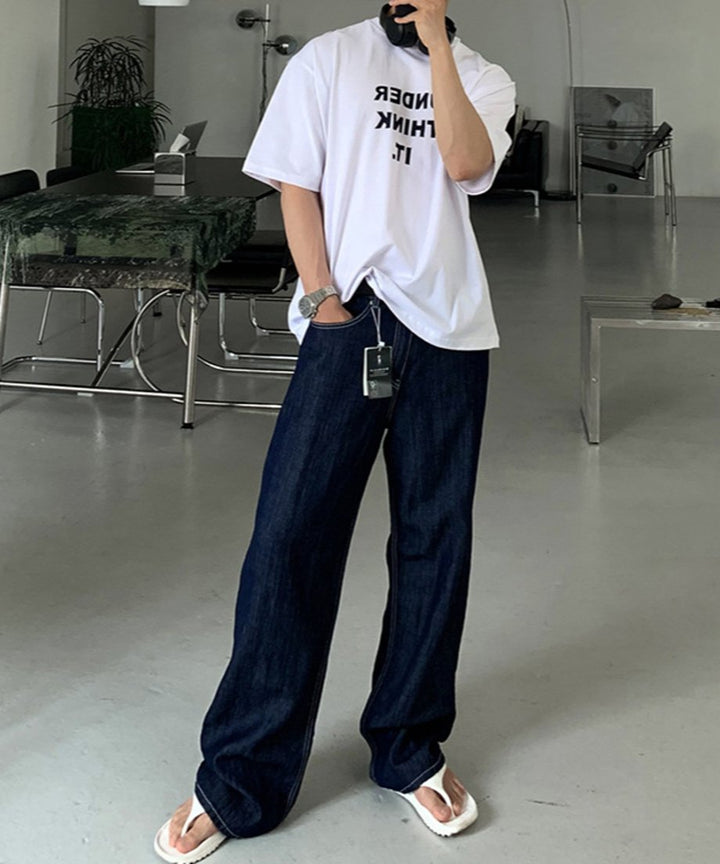 ホワイトのロゴTシャツにインディゴデニム、白いトングサンダルを合わせた男性が、広々としたモダンな室内スタジオに立っている全身スタイリングカット。首元には黒のヘッドフォンを着用。