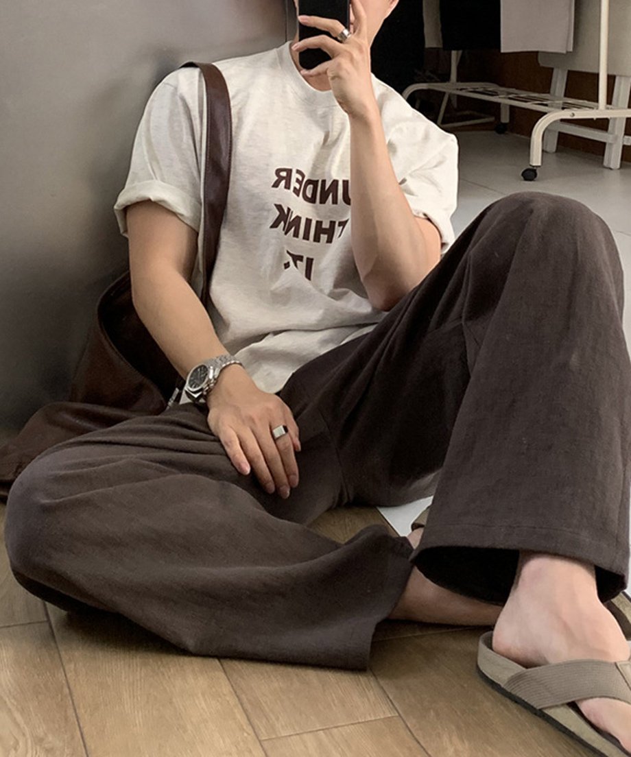 フラワーグレーのロゴTシャツを着用し、ダークブラウンのワイドパンツにベージュのトングサンダルを合わせた男性が床にラフに座り込んでいる、リラックス感漂う鏡越しの自撮りカット。