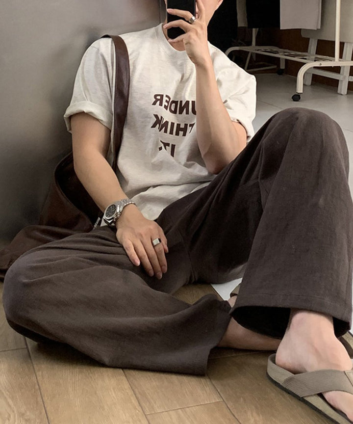 フラワーグレーのロゴTシャツを着用し、ダークブラウンのワイドパンツにベージュのトングサンダルを合わせた男性が床にラフに座り込んでいる、リラックス感漂う鏡越しの自撮りカット。