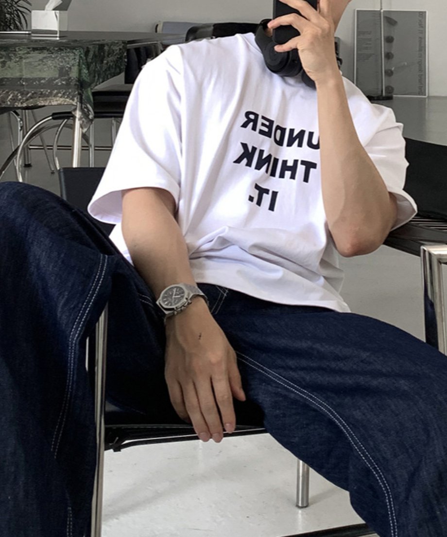 フロントに「UNDER THINK IT.」と反転・配置されたロゴがあるホワイトのTシャツを着用し、インディゴのワイドデニムを穿いてデザインチェアに腰掛ける男性のミドルショット。メタルケースの腕時計が輝く手元。
