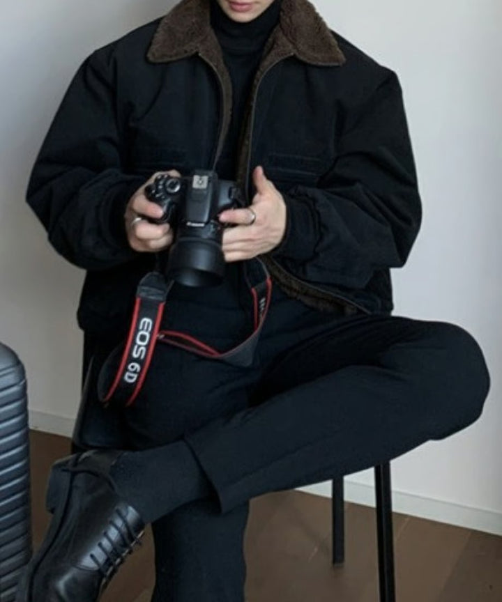 椅子に座り、一眼レフカメラ（Canon EOS 6D）を両手で持つ男性のスタイリングカット。黒のタートルネック、黒のスラックス、レザーシューズを合わせたクリーンなモノトーンコーデに、ブルゾンのブラウンボア襟が知的なアクセントを添えている。