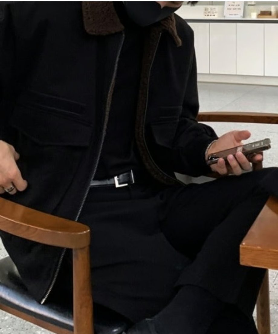 カフェの椅子に腰掛け、スマホを操作する男性のウエストアップカット。フロントジップを開けたブルゾンから覗く、黒のインナーとレザーベルト、シルバーバックルの組み合わせがシックな印象。