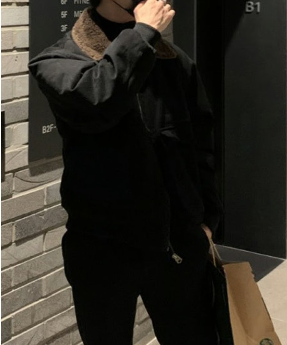 案内板の前で少し身体を傾けて立つ男性の斜めからのカット。ブルゾンのフロントジップを少し下げ、襟元のボアを見せ、手でマスクを触れている画像。