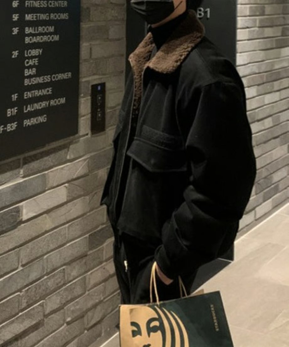 案内板の前で少し身体を傾けて立つ男性の斜めからのカット。ブルゾンのフロントジップを少し下げ、襟元のボアを見せつつ、袖口のしっかりとしたリブのホールド感が伝わるショット。