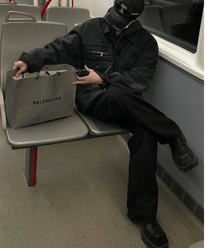 電車のシートに深く腰掛け、足を組んで座る男性のフルスタイリングカット。キャップとサングラスを着用し、手元にはバレンシアガ（BALENCIAGA）のショップバッグを添えた、ラグジュアリーストリートな世界観を体現した1枚。