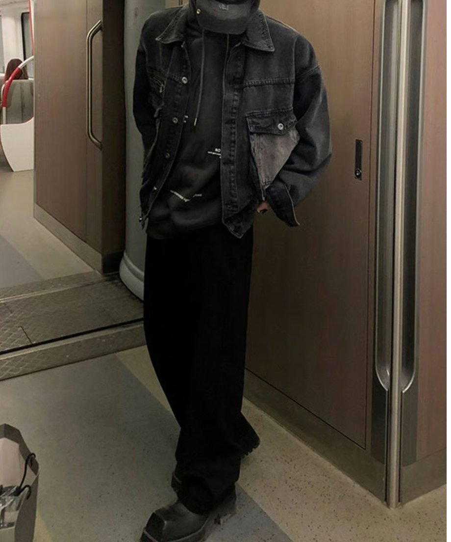 電車の連結部付近に立つ男性の全身スタイリングカット。オーバーサイズのデニムジャケットに、ワイドなブラックパンツと重厚なスクエアトゥのブーツを合わせた都会的なオールブラックコーデ。