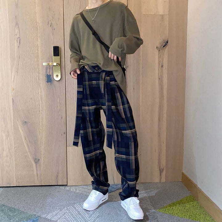 木目のドアの前で、ネイビーのチェック柄ワイドパンツにカーキの長袖カットソー、白のスニーカーを合わせた男性の全身正面カット。共布のベルトがフロントでルーズに垂れ下がっています。