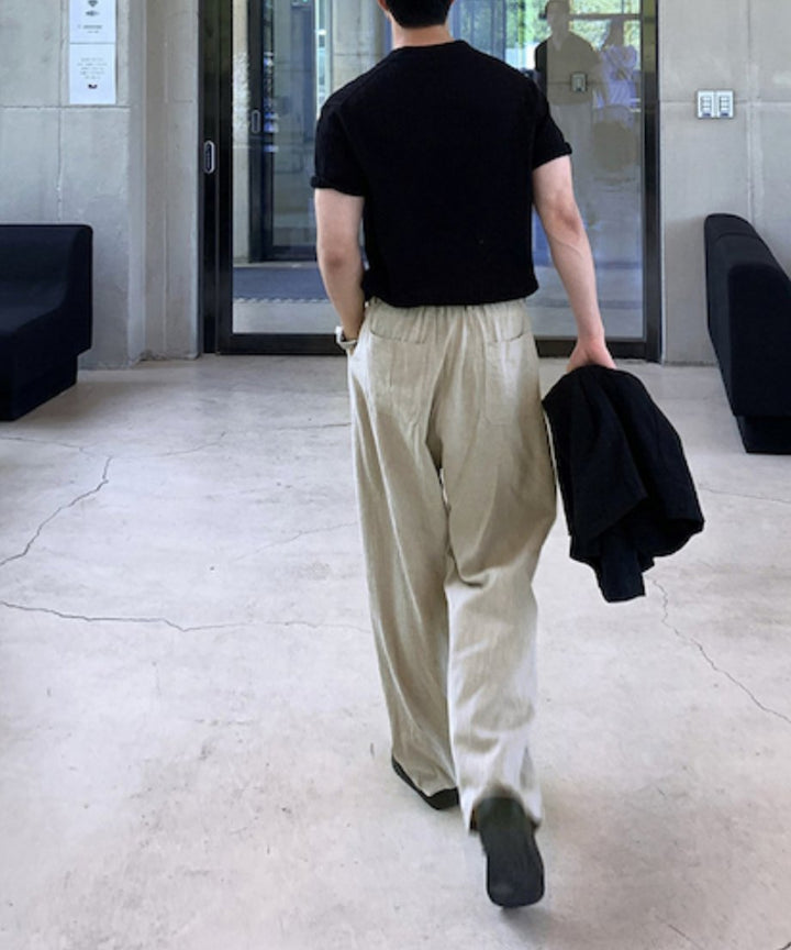 黒の半袖ニットにライトベージュのリネン混ワイドパンツを合わせ、黒いジャケットを手に持ってカフェの通路を歩き去る男性の後ろ姿。