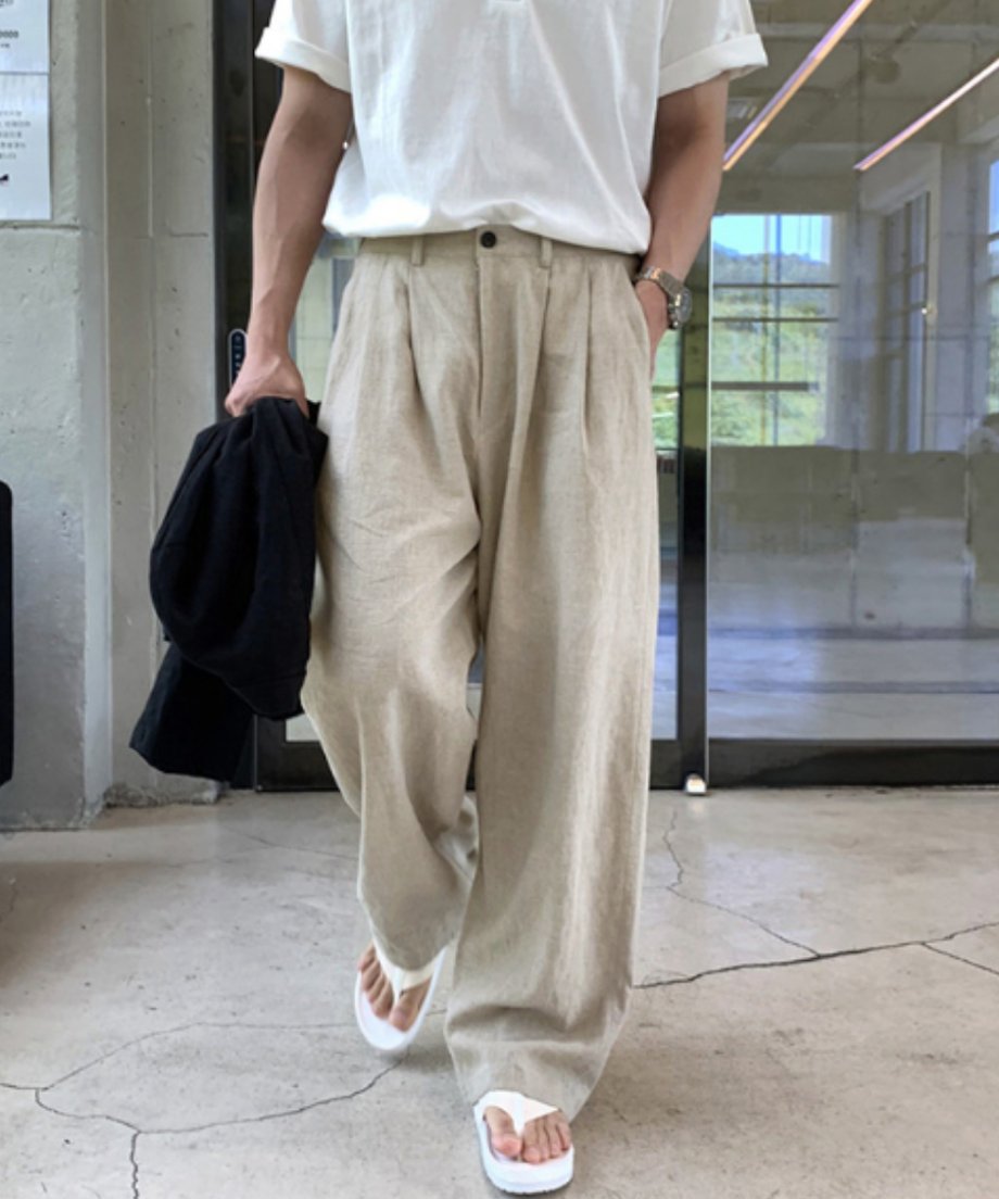 ベージュのリネンタックパンツに白いTシャツと白いトングサンダルを合わせ、黒いジャケットを手に持って歩く男性の正面コーディネート。