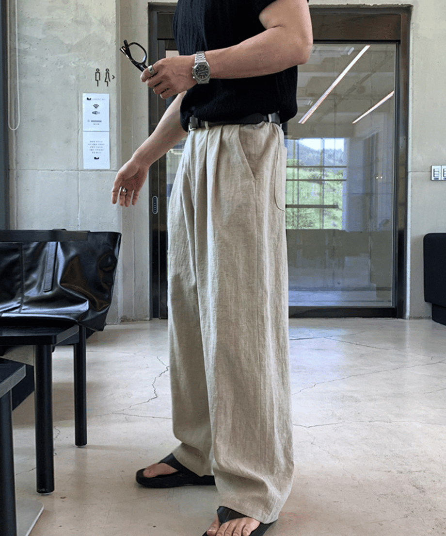 ベージュのワイドパンツに黒のレザーベルトと腕時計を合わせ、手にサングラスを持った男性のサイドフロントカット。