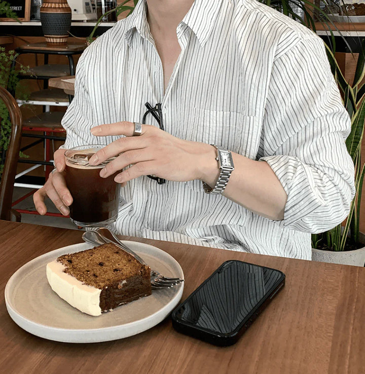 カフェのウッドテーブル席で、白いストライプシャツを着用してアイスコーヒーのグラスを持つ男性の手元。左腕にはスクエア型のメタルバンド腕時計、人差し指には太めのシルバーリングが光っています。テーブルにはケーキ皿が置かれています。